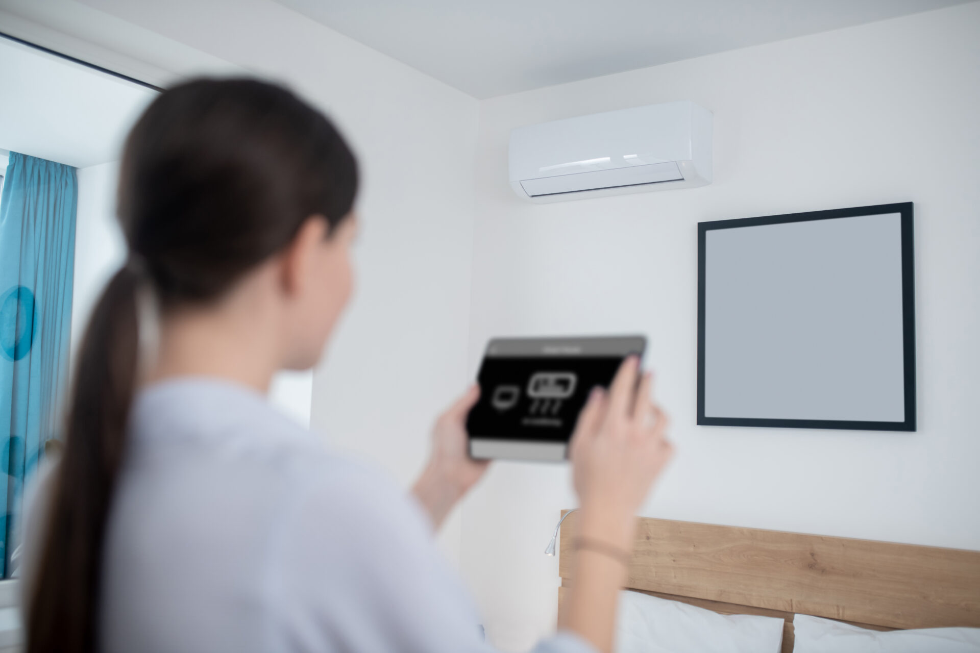 Woman turning on an air conditioner using a tablet Back view of a dark-haired lady with the electronic device in the hands standing before the wall-mounted air conditioning unit